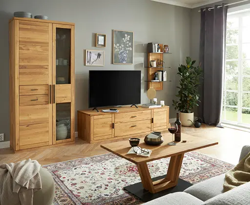 Wohnzimmer mit massiven Holzmöbeln, Fernseher, Teppich und großen Fenstern.